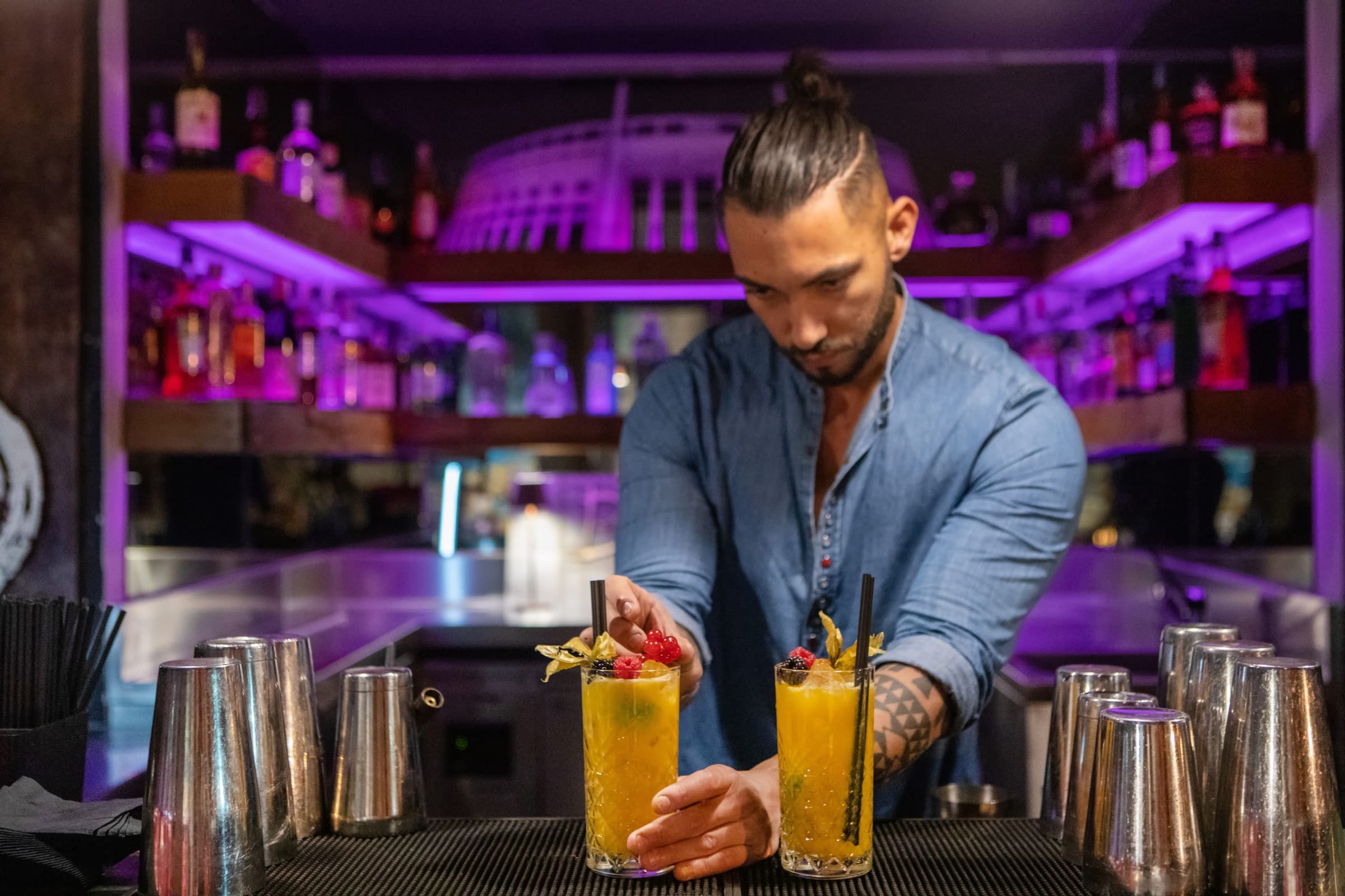 Bartender con camicia blu serve due cocktail con frutta al bancone