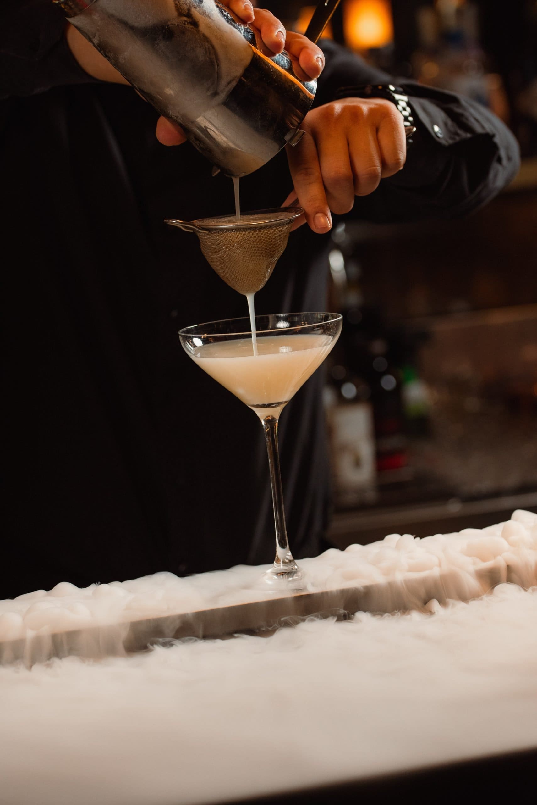 Bartender preparing a cocktail with a shaker and strainer over a martini glass.